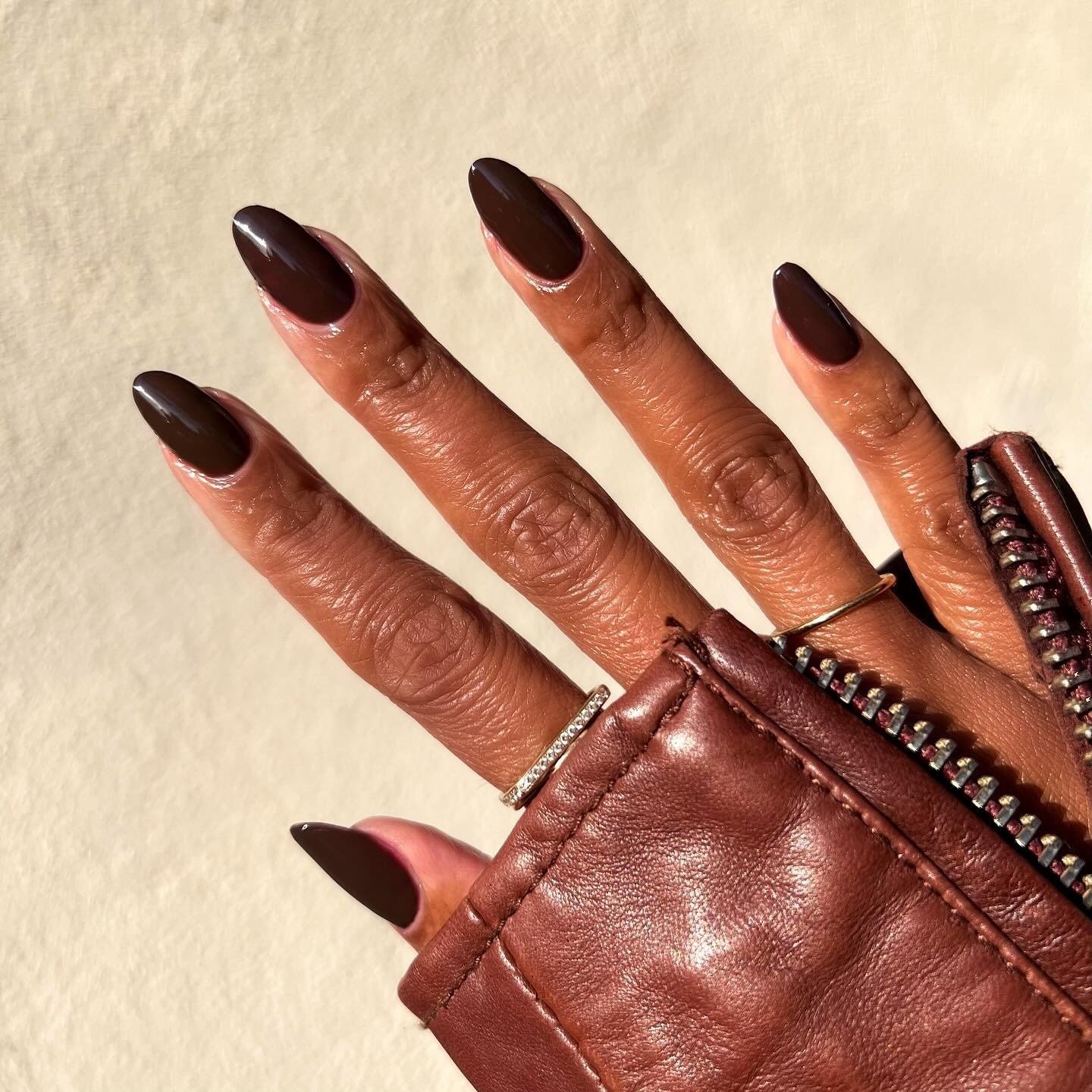 Unhas longas com esmaltes em alta na cor marrom chocolate brilham junto a jaqueta de couro vinho e anéis delicados,