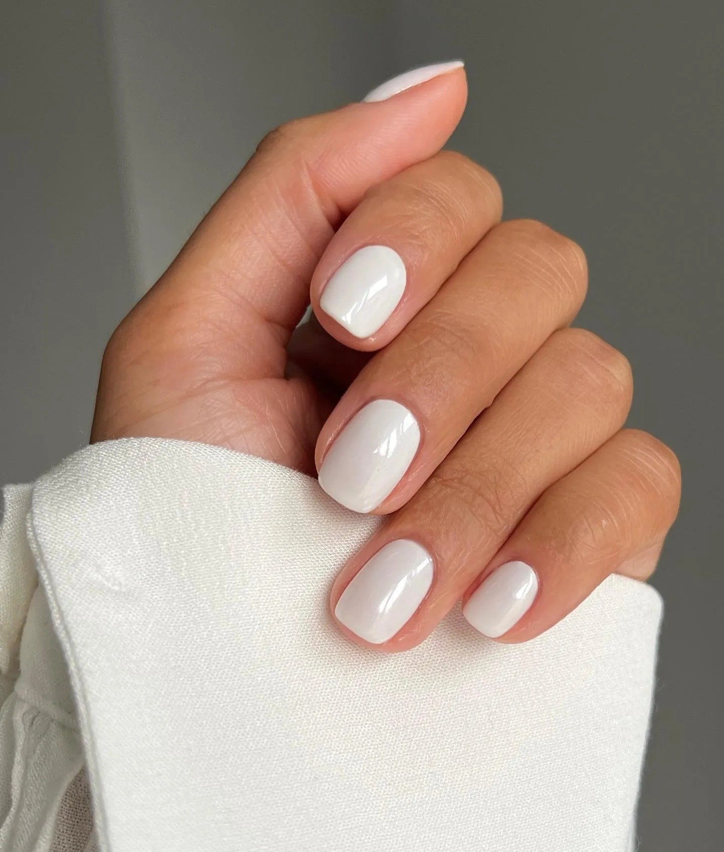 Esmaltes em alta com acabamento branco perolado realçam unhas curtas e arredondadas, combinando com tecido branco suave para