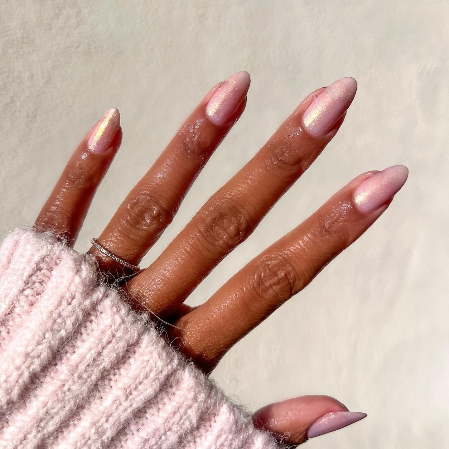 Mão com esmaltes em alta em tom rosa perolado, unhas alongadas em formato amêndoa, combinando com suéter rosa de tricô macio.