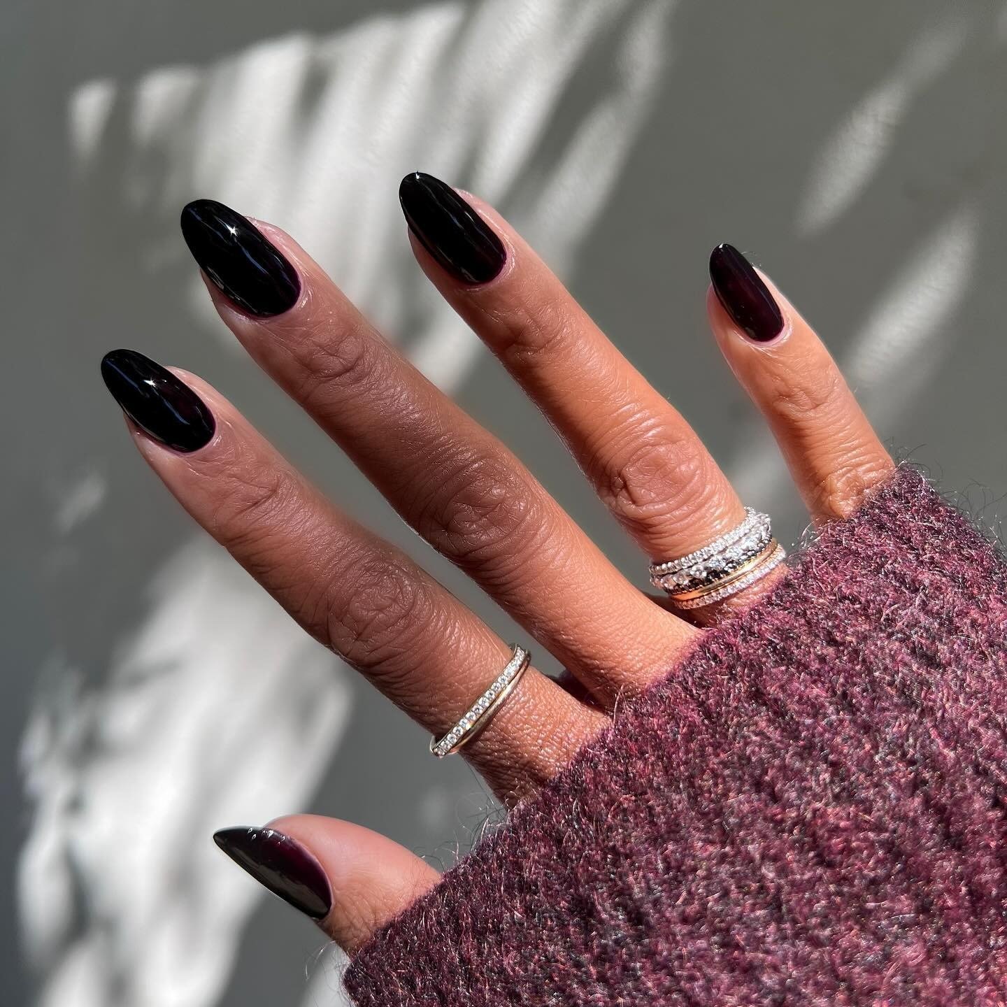 Mão com esmaltes em alta preto brilhante, anéis delicados e suéter roxo de tricô, em luz natural suave e elegante.