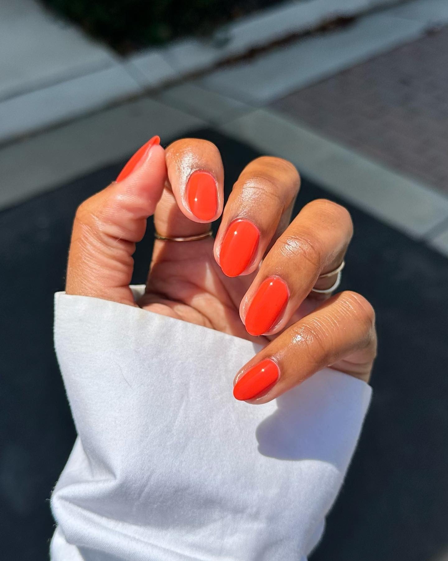 Mão com esmaltes em alta laranja vibrante, unhas ovais bem cuidadas e manga branca de tecido leve ao sol.