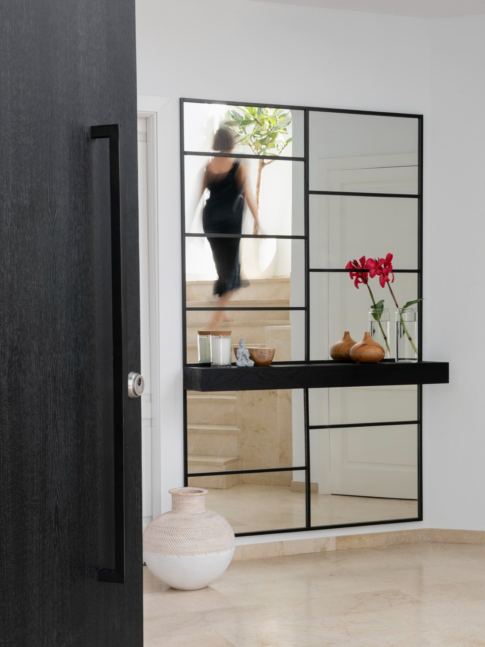 Espelho em casa com estrutura preta e prateleira minimalista exibe decoração com vasos de madeira e flores vermelhas