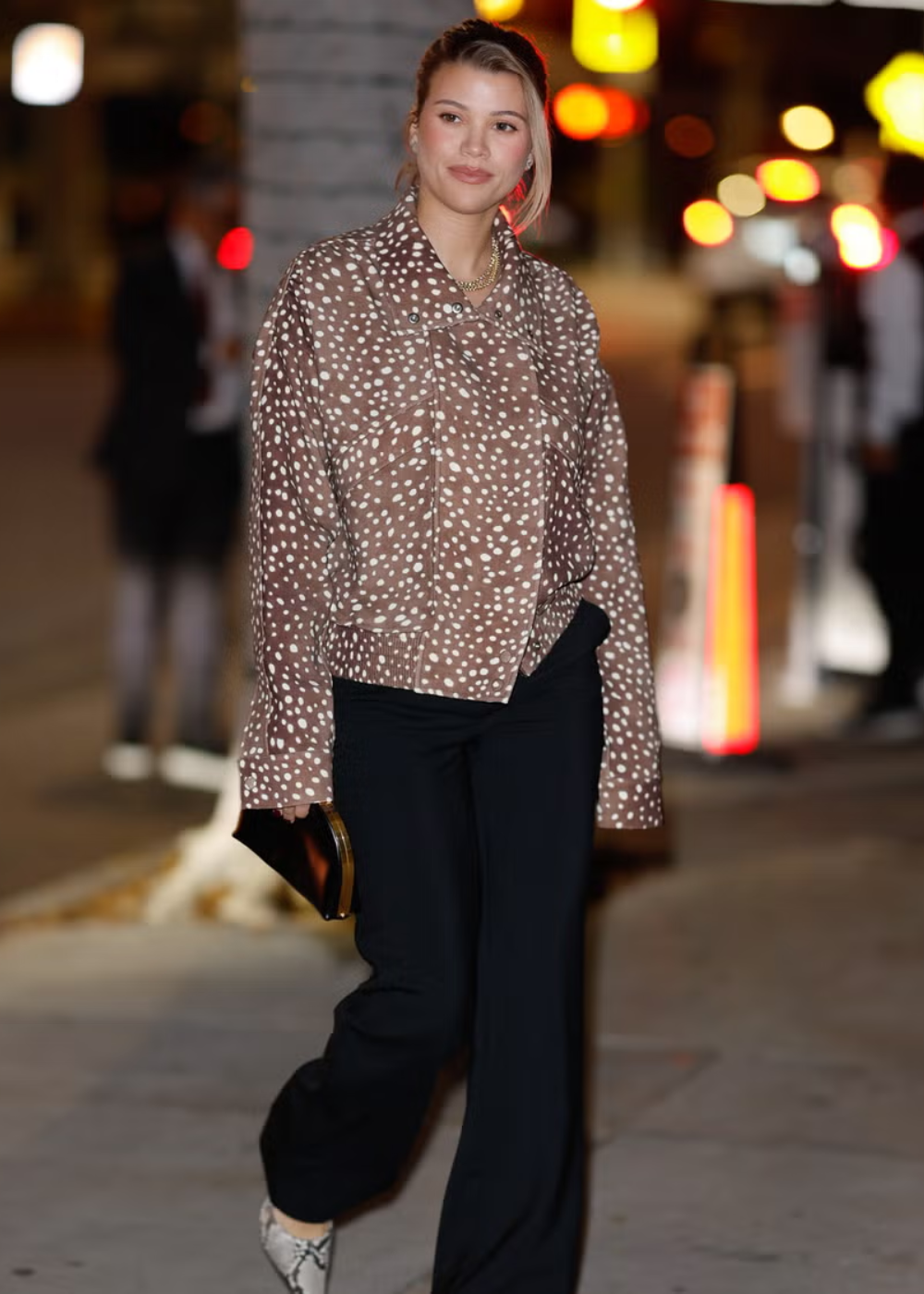 Modelo caminha com jaqueta oversized de estampa de cervo marrom com bolinhas brancas, combinada a calça preta reta e scarpin.