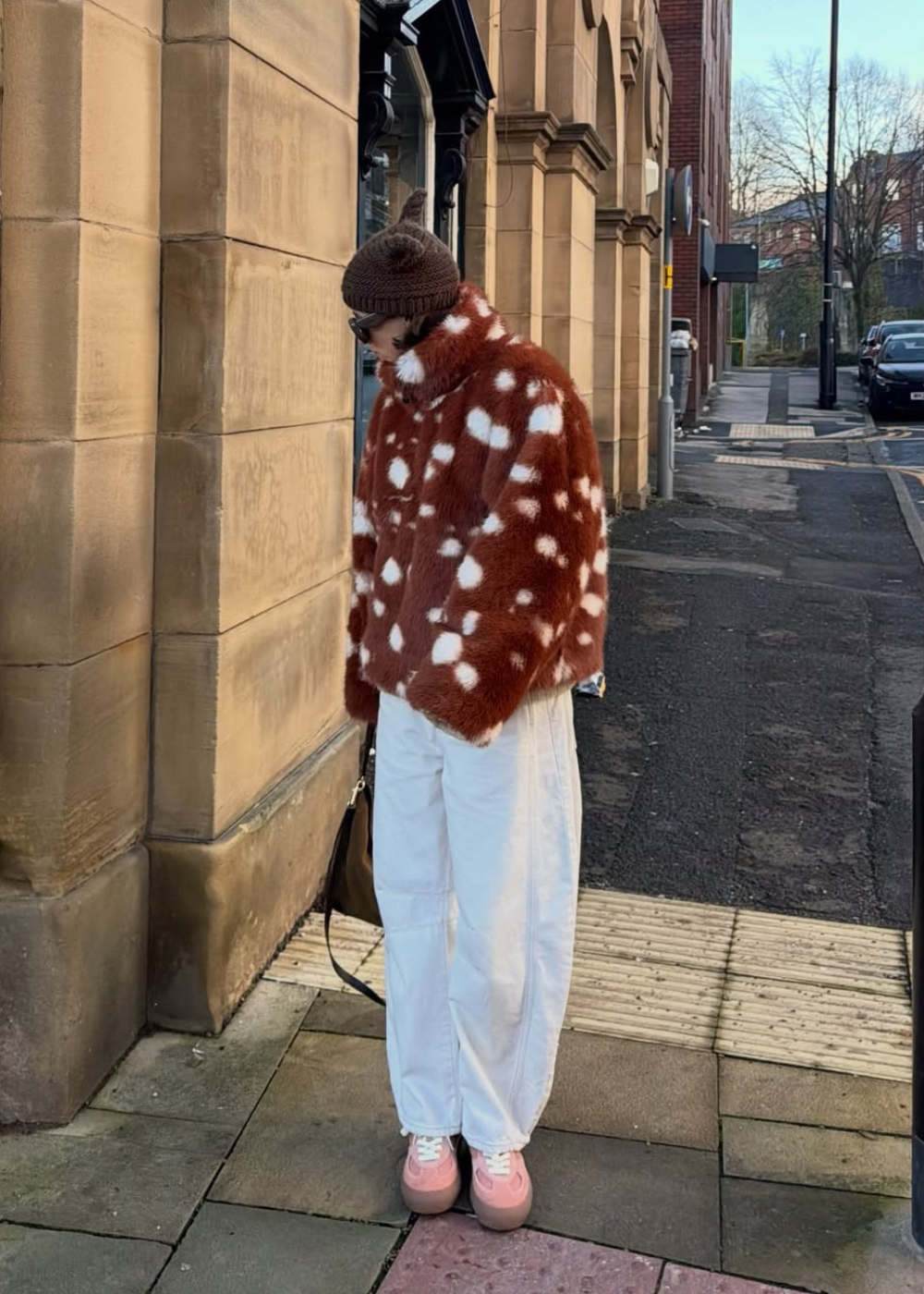 Modelo posa com casaco peludo de estampa de cervo em tons terrosos e branco, calça branca ampla e tênis rosa.