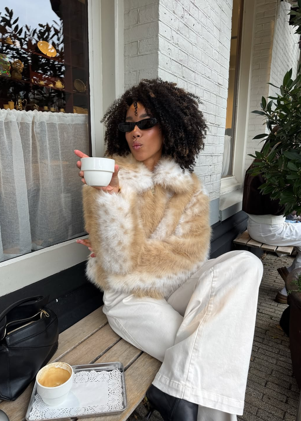 Modelo posa sentada com jaqueta de pelúcia em estampa de cervo bege e branca, calça branca ampla e botas pretas.