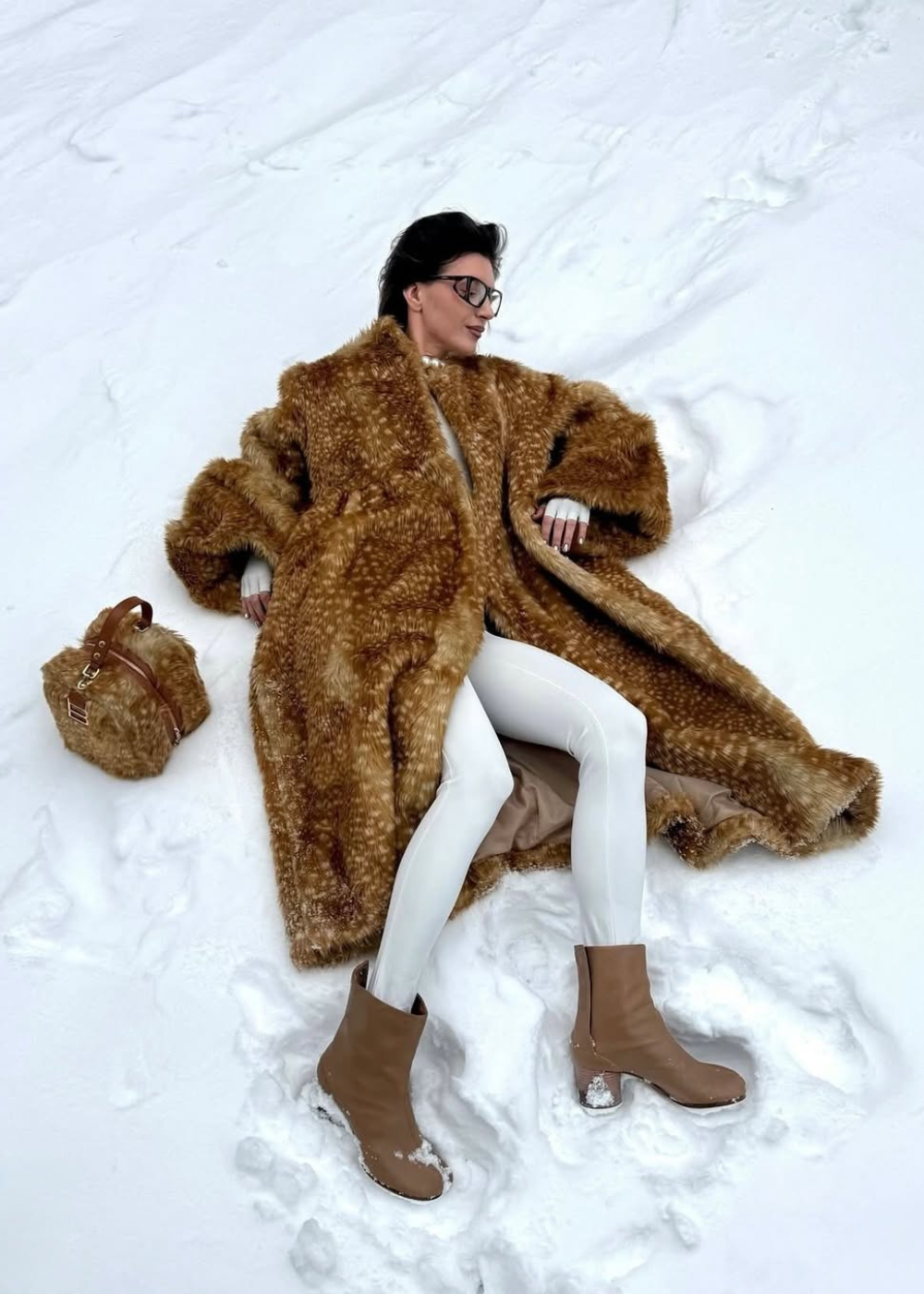 Modelo posa deitada na neve com casaco oversized peludo de estampa de cervo em tons terrosos, legging branca e botas bege.