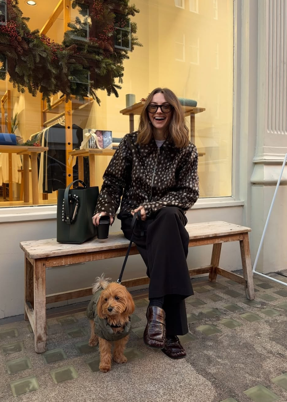 Mulher senta sorrindo com jaqueta de estampa de cervo em tons terrosos, calça preta e mocassim marrom.