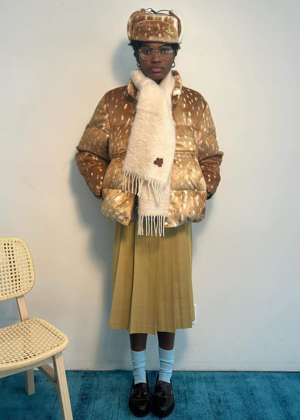 Modelo posa com jaqueta e chapéu acolchoados em estampa de cervo marrom com branco, saia plissada mostarda, cachecol creme.