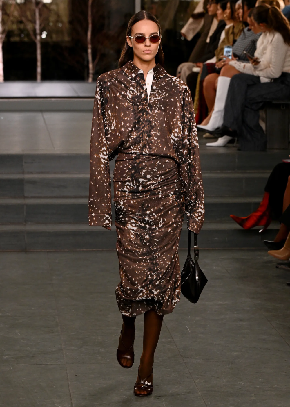 Modelo caminha com postura confiante usando vestido de cetim marrom com estampa de cervo, mangas longas e saia drapeada.