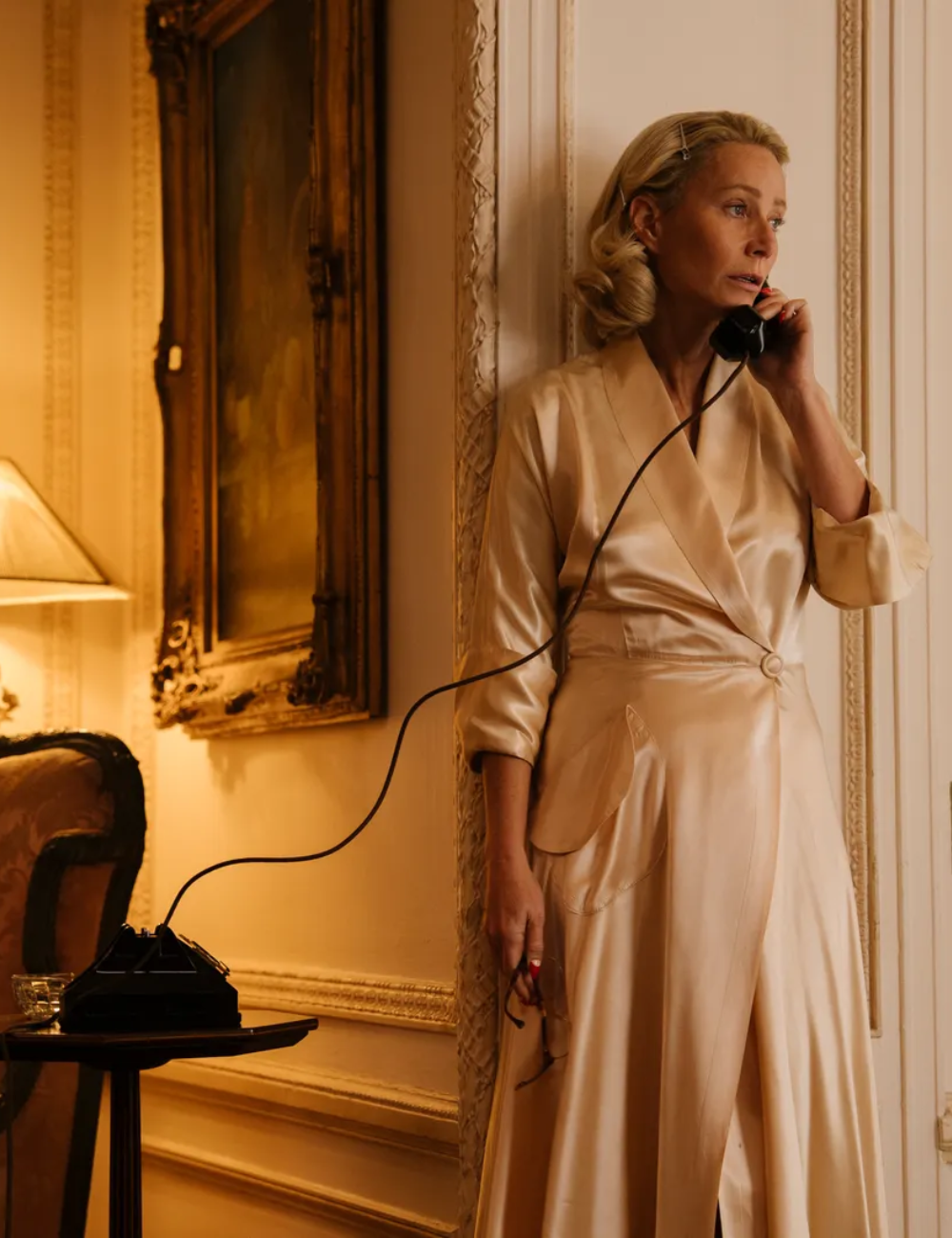 Mulher posa pensativa ao telefone em ambiente clássico, vestindo vestido longo Marty Supreme de cetim bege.