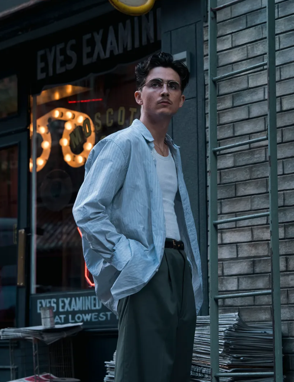 Modelo posa com camisa azul clara oversized de algodão leve, regata branca e calça verde escura de alfaiataria.