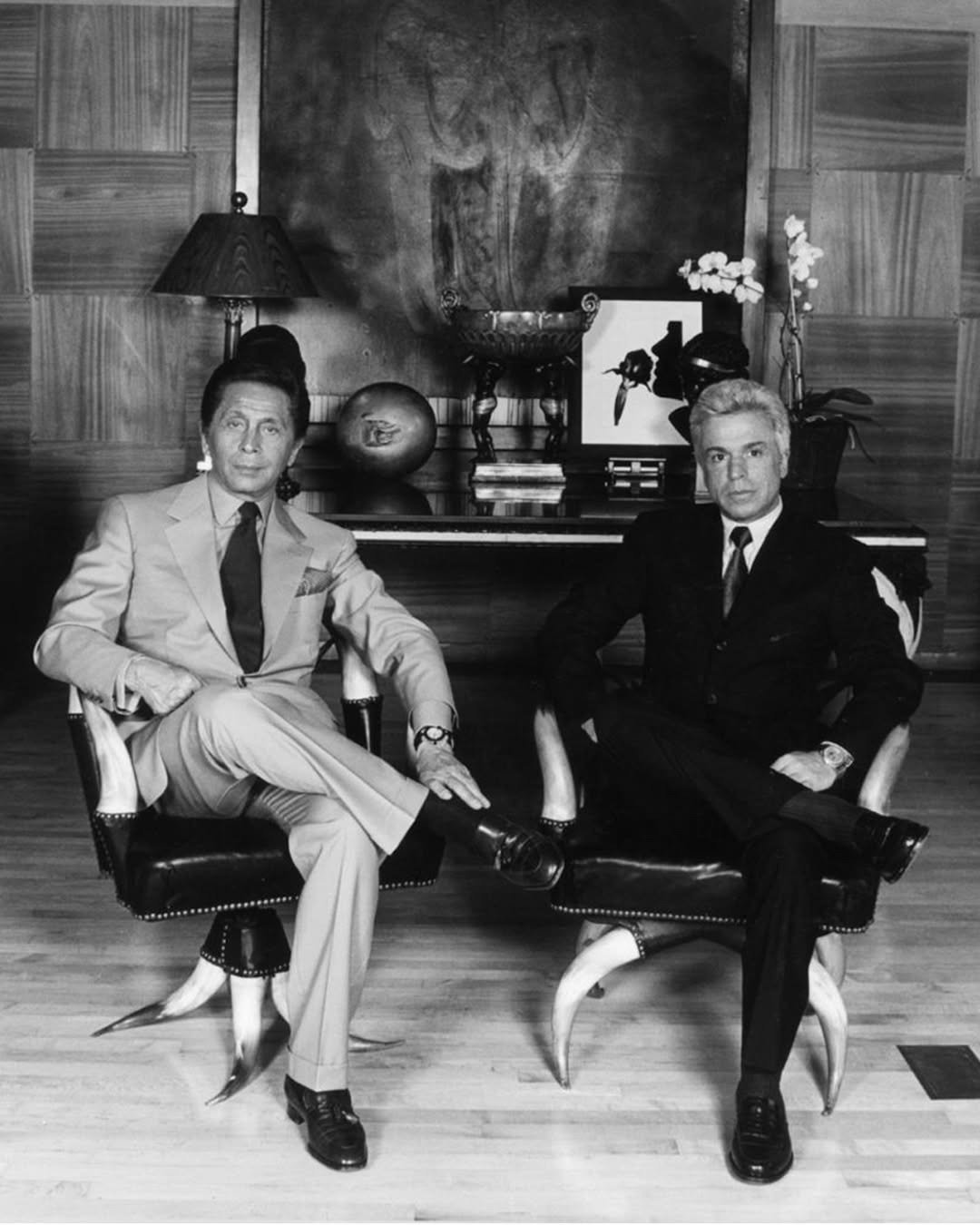 Dois homens posam sentados com postura confiante em cadeiras de design único, vestindo ternos elegantes Valentino, um bege