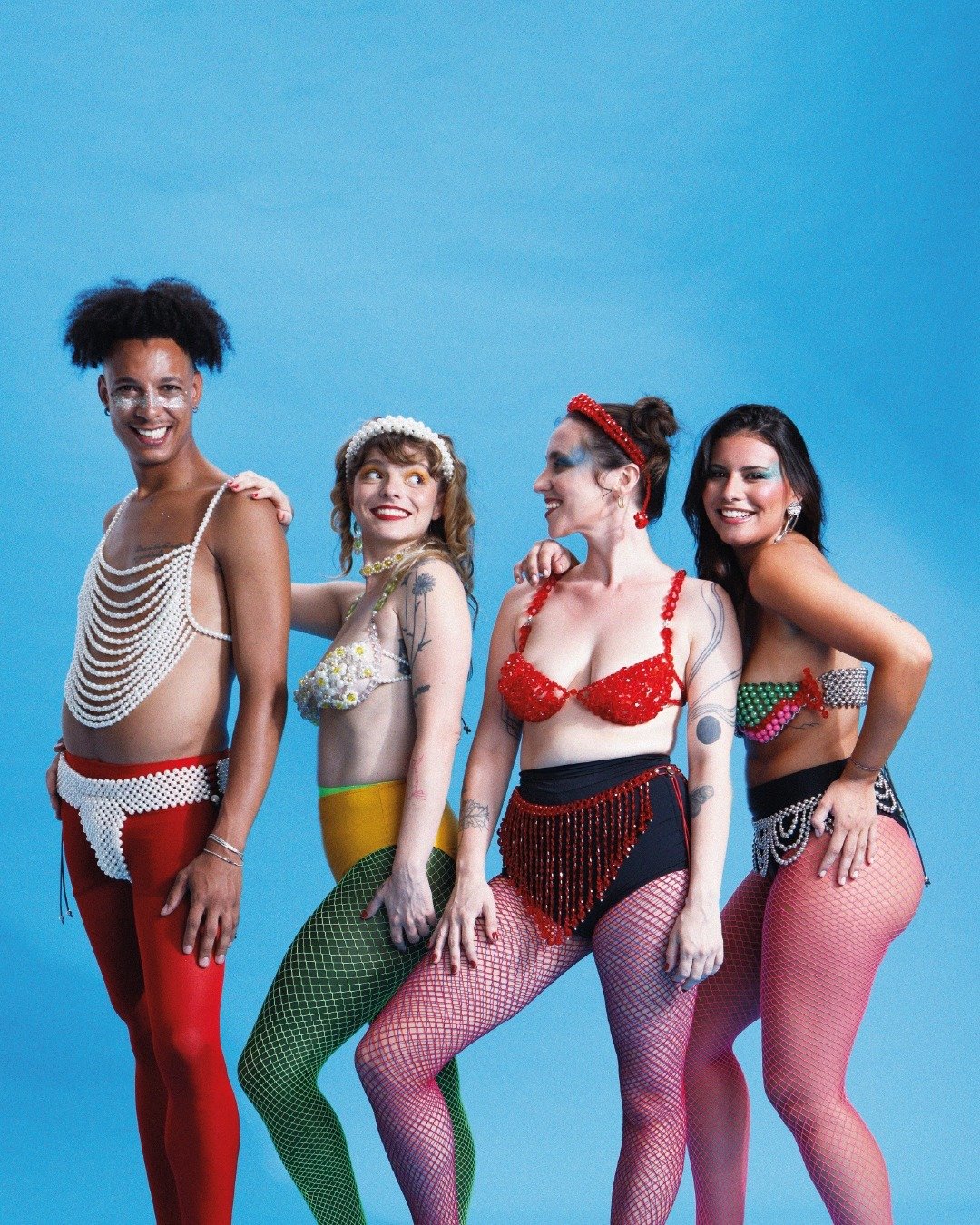 Quatro modelos posam sorrindo com acessórios carnavalescos em pérolas, miçangas e meia arrastão colorida, celebrando a