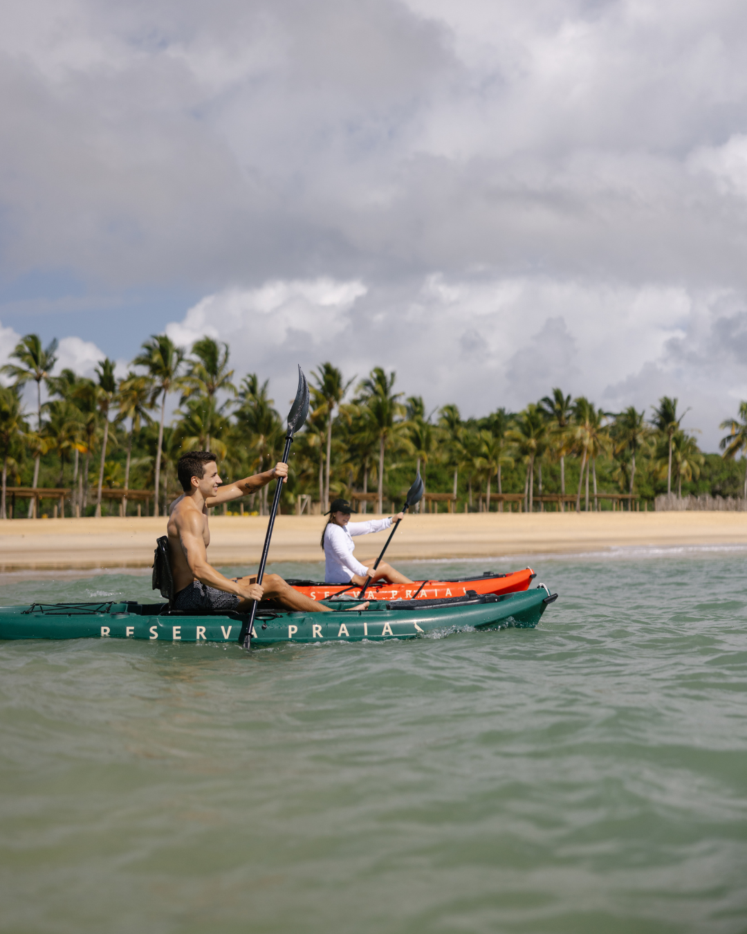 Casal remando em caiaques coloridos na praia, clima ativo e descontraído no guia do verão brasileiro