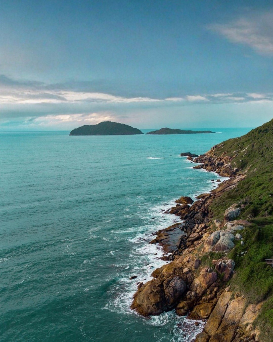 Costa rochosa com mar azul-turquesa sob céu nublado, cenário natural que inspira o guia do verão brasileiro.
