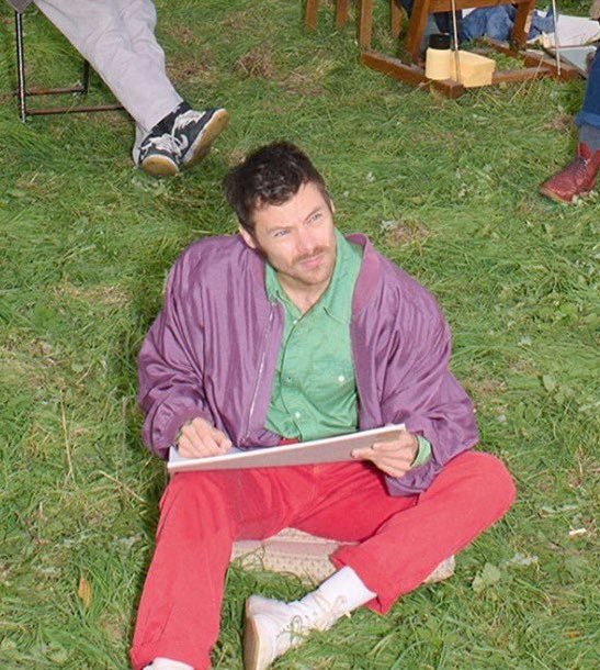 Homem sentado na grama veste jaqueta lilás, camisa verde e calça vermelha, destacando tênis polêmico branco com detalhes