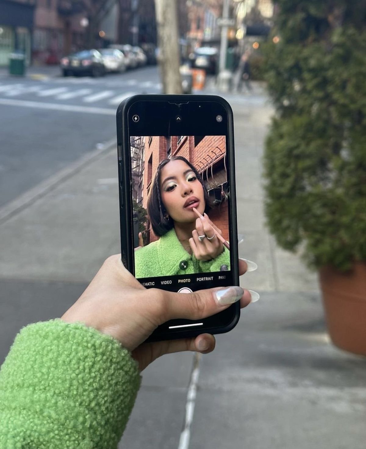 Modelo posa para selfie no Instagram usando casaco verde de textura felpuda e maquiagem suave, em cenário urbano.