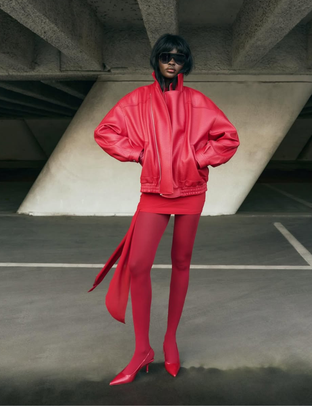 Modelo posa com jaqueta oversized de couro vermelho, minissaia com faixa fluida e meia-calça combinando.