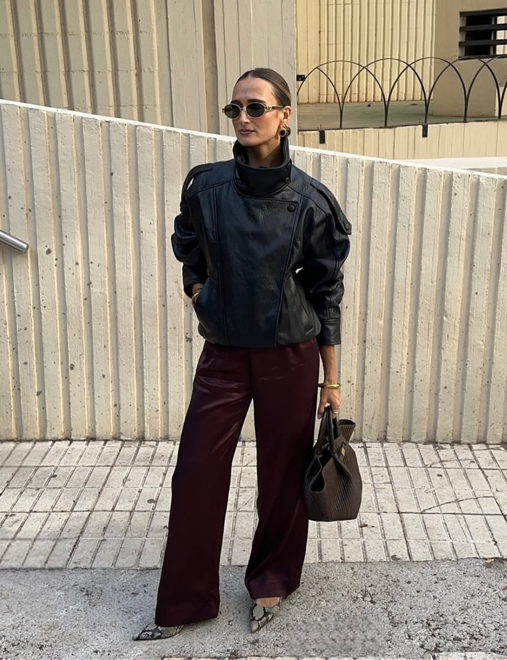 Modelo posa com jaqueta de couro preta oversized com ombros estruturados, calça vinho ampla e sapato de estampa animal.