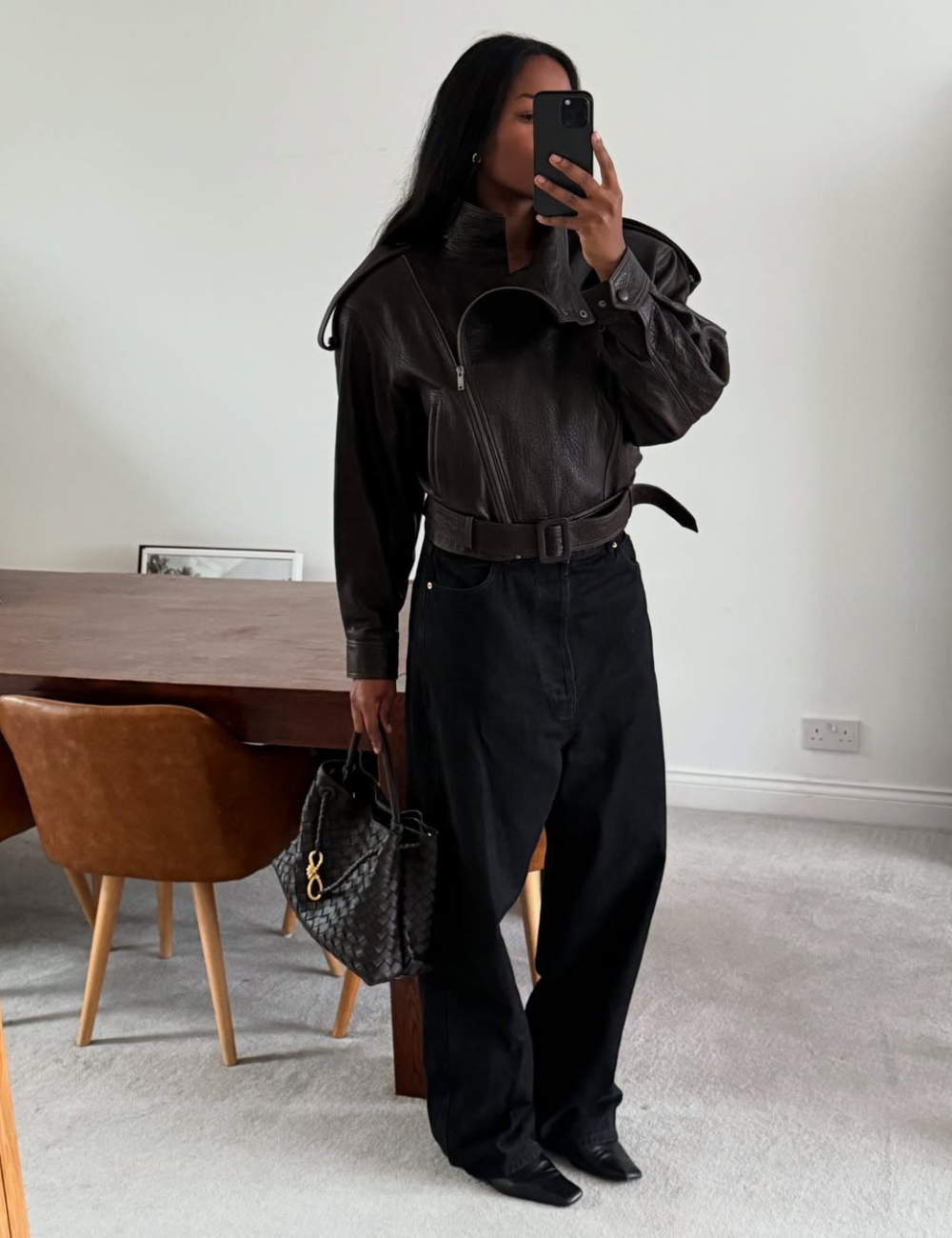 Modelo posa segurando celular e bolsa, vestindo jaqueta de couro oversized com ombreiras e calça preta ampla.