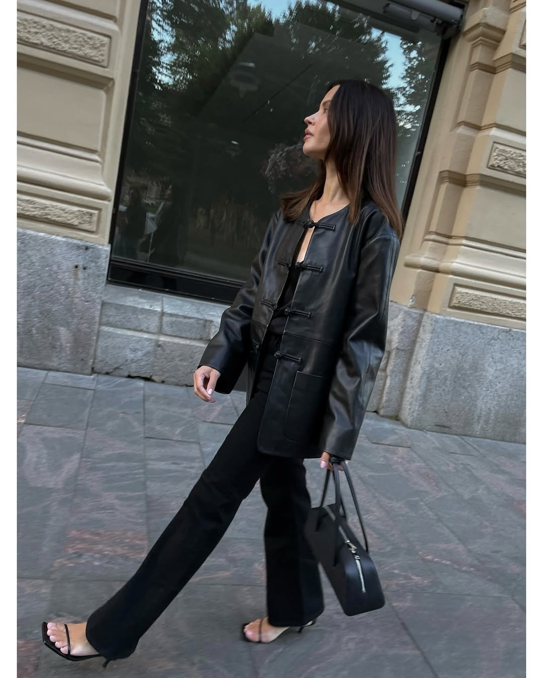 Modelo caminha confiante com jaqueta de couro preta com nó chinês, calça reta, sandálias delicadas e bolsa estruturada preta.