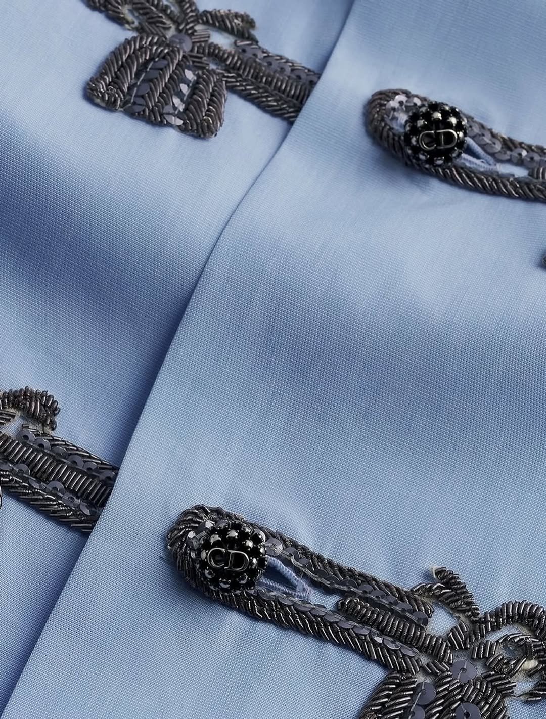 Detalhe de nó chinês em tecido azul claro com bordados pretos e botões texturizados, evidenciando sofisticação e delicadeza.