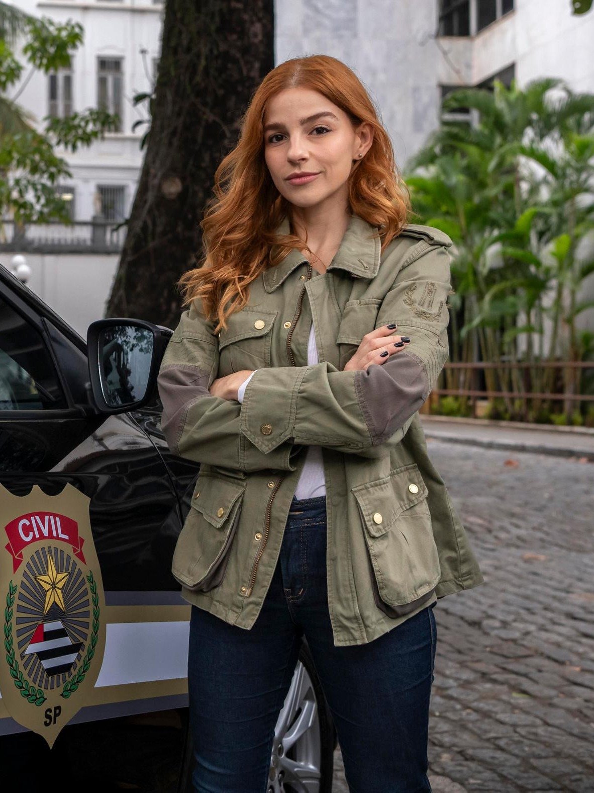 Lorena e Juquinha aparecem com jaqueta utilitária verde oliva de bolsos frontais, jeans escuro e postura confiante diante de