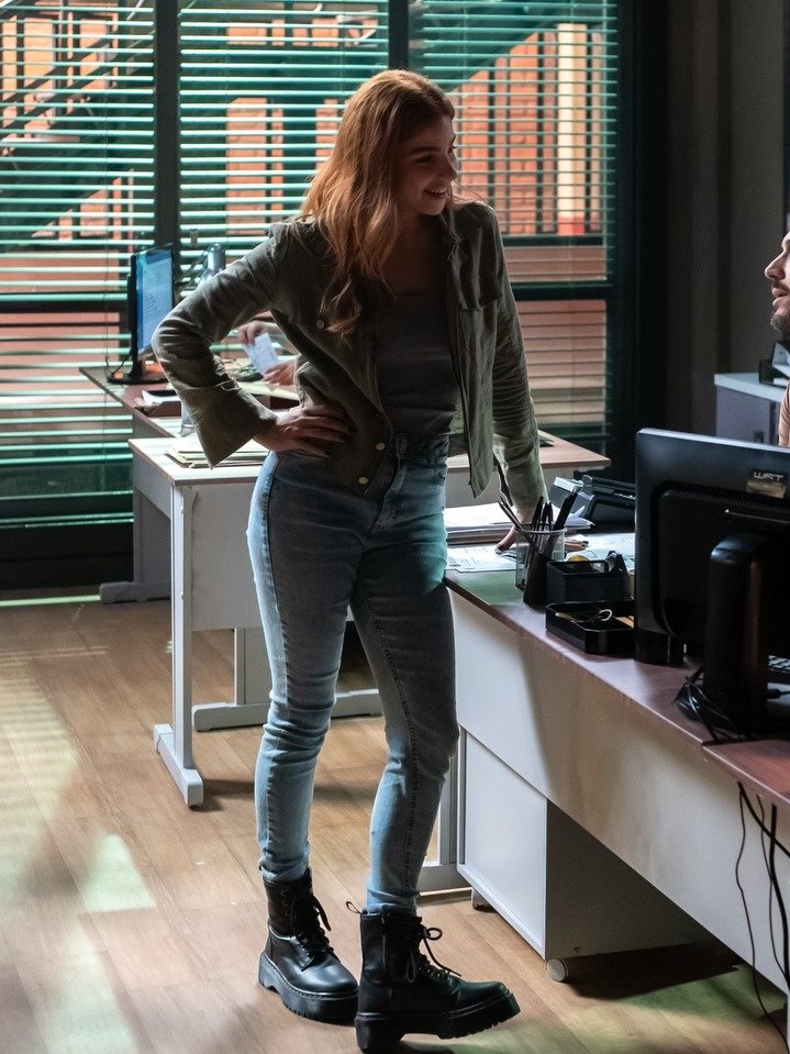 Lorena usa jaqueta verde militar, regata cinza, jeans skinny azul claro e coturno preto, posando confiante em escritório