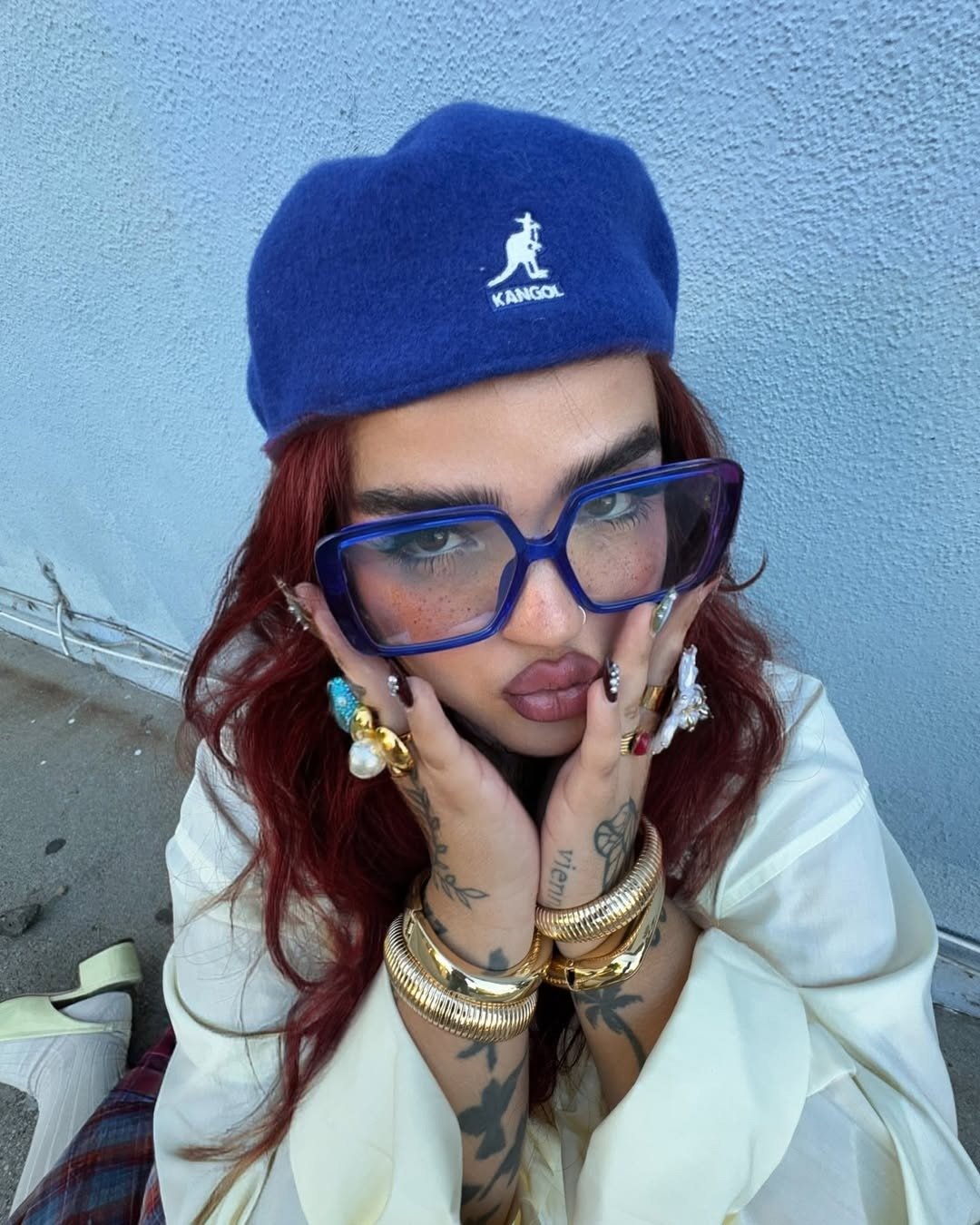 Modelo posa com boina Kangol azul de lã, óculos oversized azuis, anéis e braceletes dourados, transmitindo estilo ousado