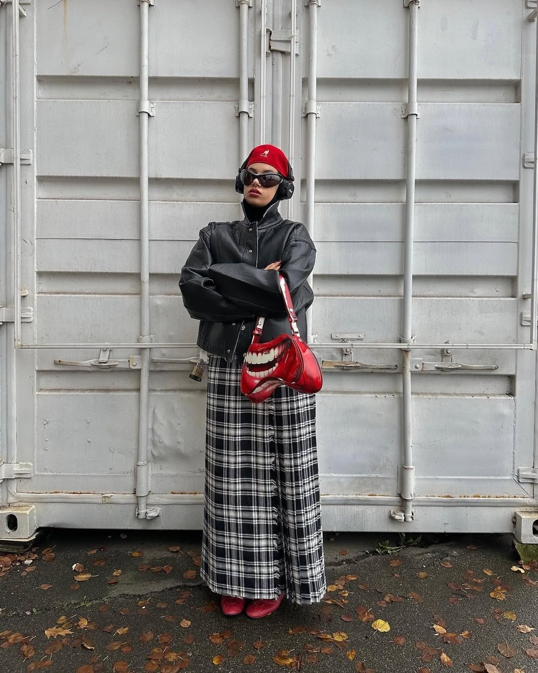 Modelo posa com boina kangol vermelha de lã, jaqueta preta de couro, saia longa xadrez e bolsa vermelha com estampa de boca