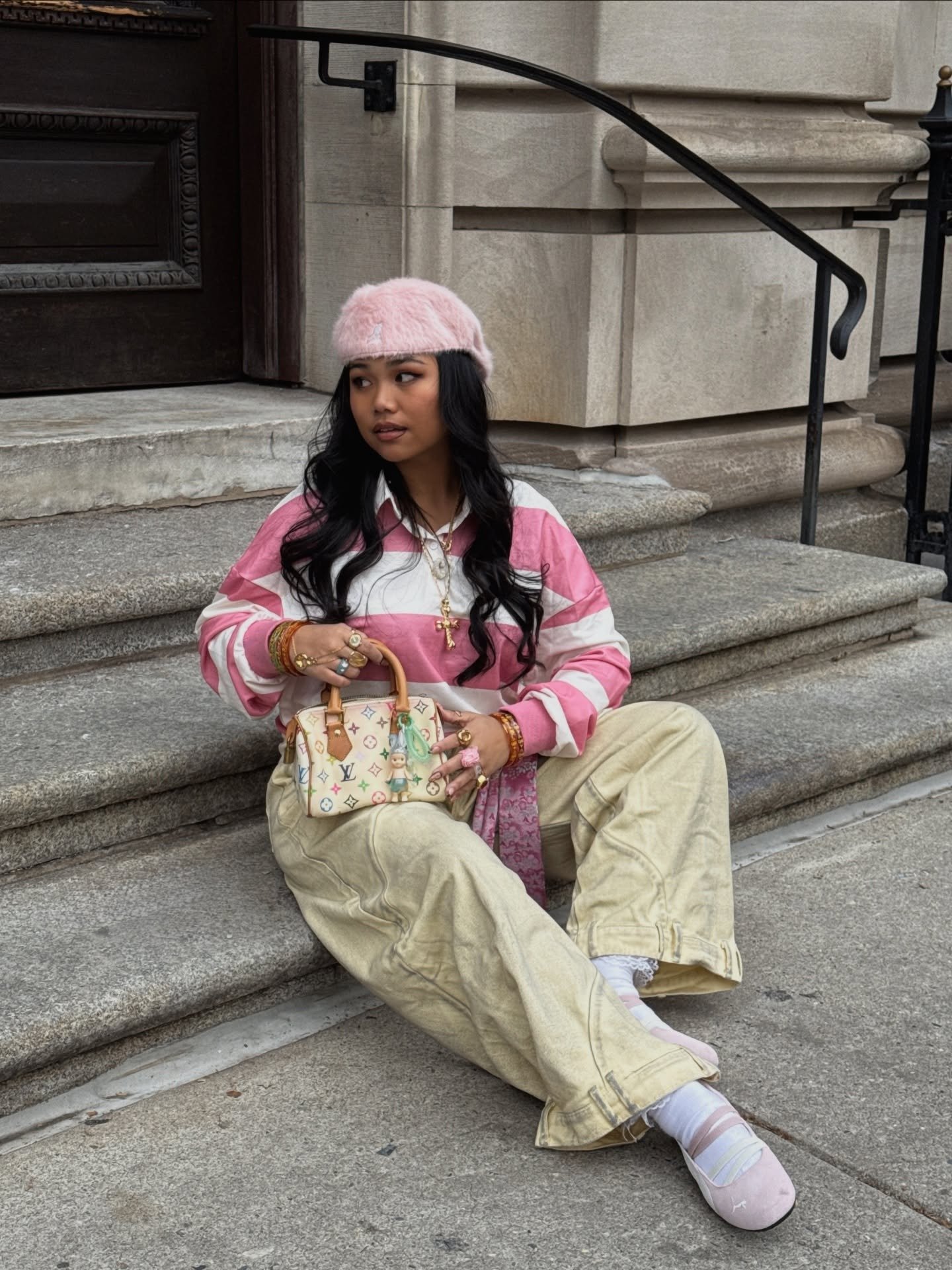 Modelo senta nas escadas com boina kangol rosa peluda, blusa listrada rosa e branca, calça ampla bege e bolsa estampada