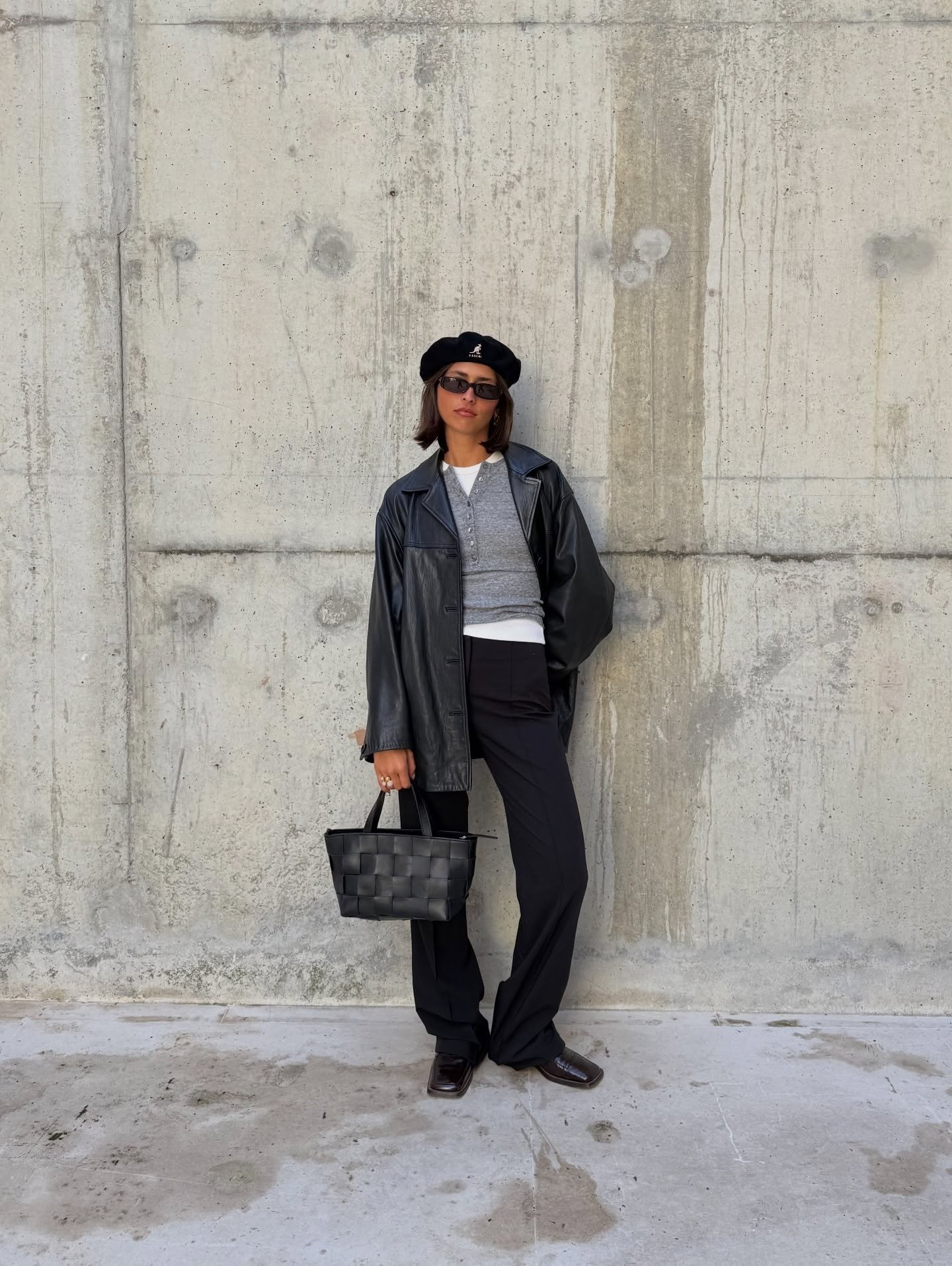 Modelo posa com boina Kangol preta de lã, jaqueta oversized de couro, blusa cinza justa, calça preta reta e bolsa de couro