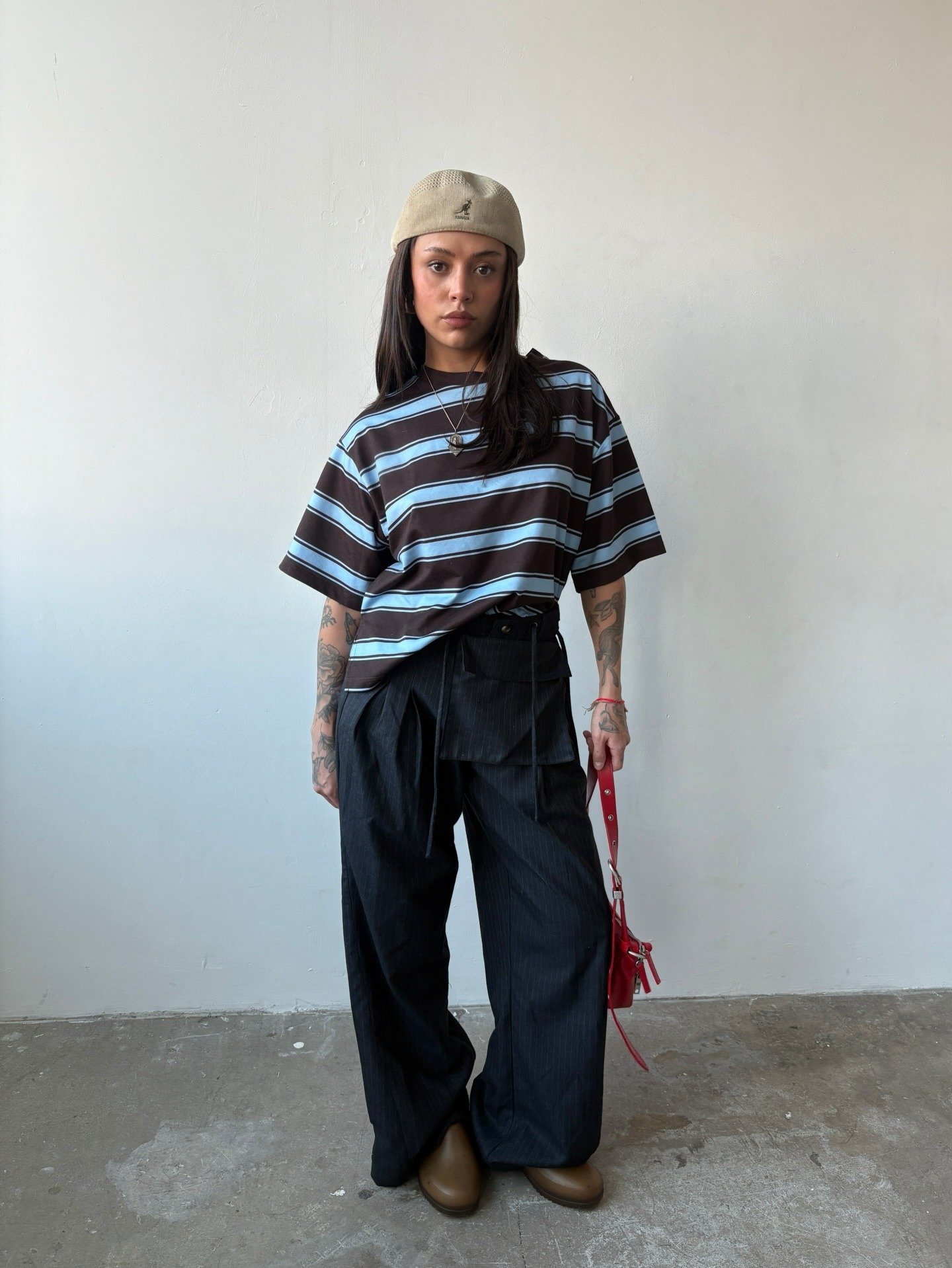 Modelo posa com boina kangol bege de tecido texturizado, camiseta listrada azul e marrom oversized, calça preta ampla