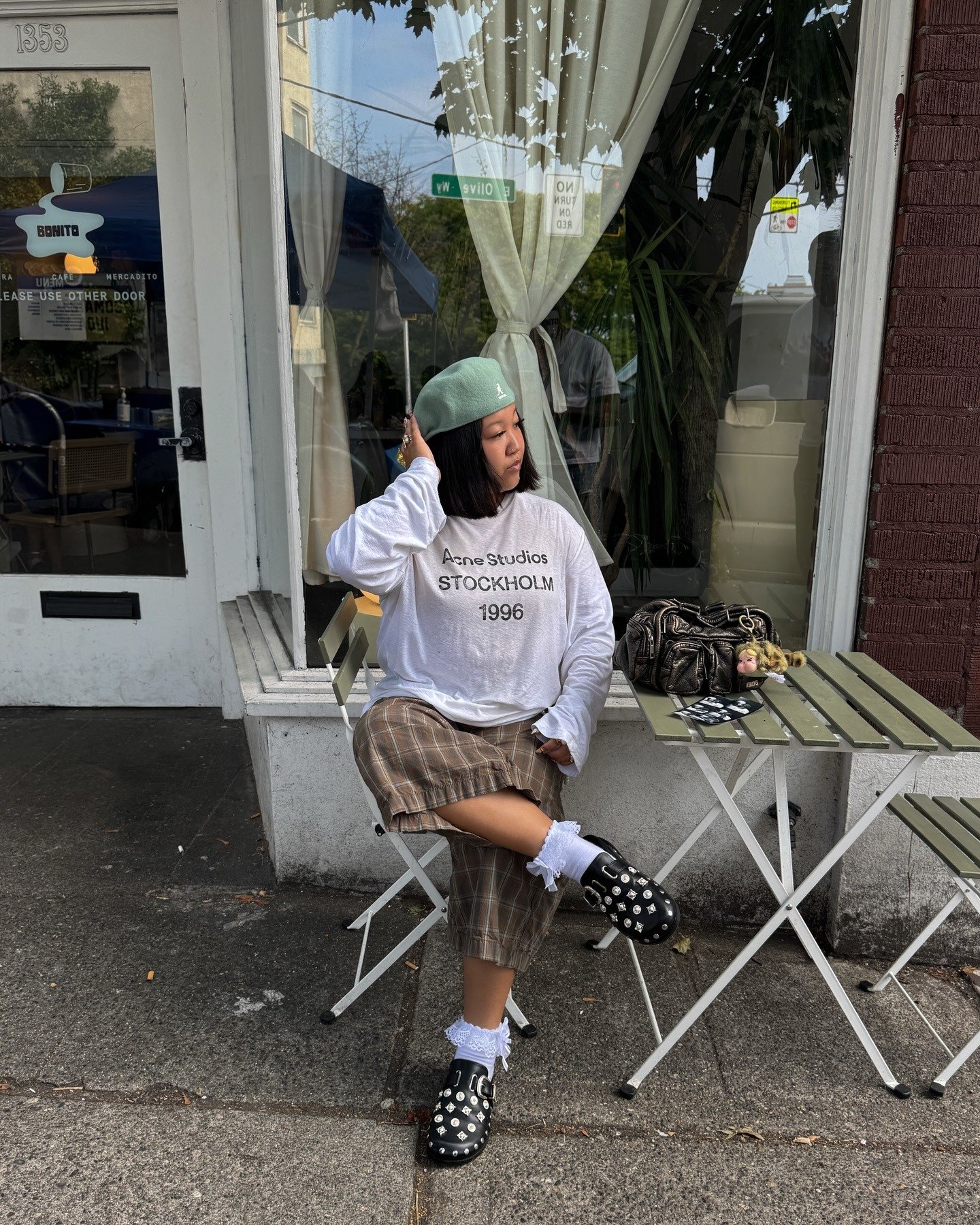 Modelo senta com postura relaxada e mão no cabelo, usando boina kangol verde menta em tecido macio, blusa branca oversized