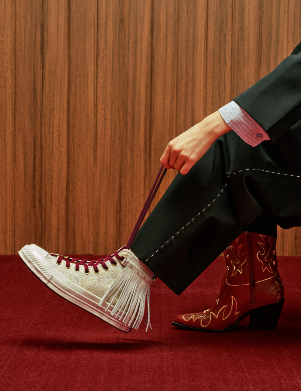 Modelo senta ajustando tênis branco com franjas e cadarço vinho, calça preta com costura aparente e bota vermelha bordada.