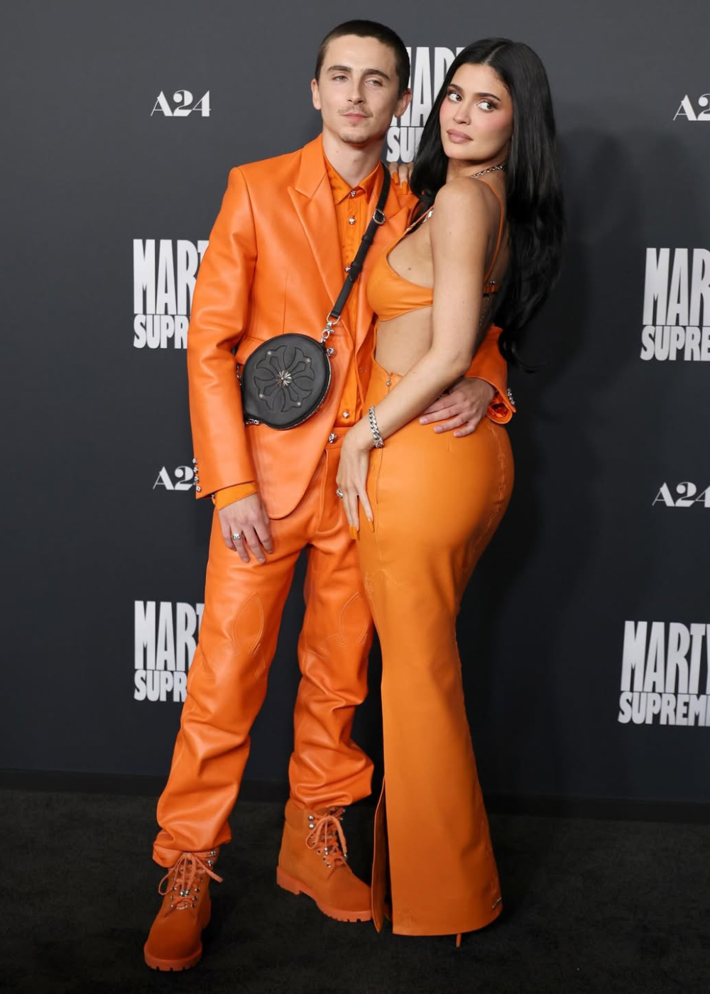Casal posa unido em looks de couro laranja vibrante, com blazer oversized, cropped e calça flare.