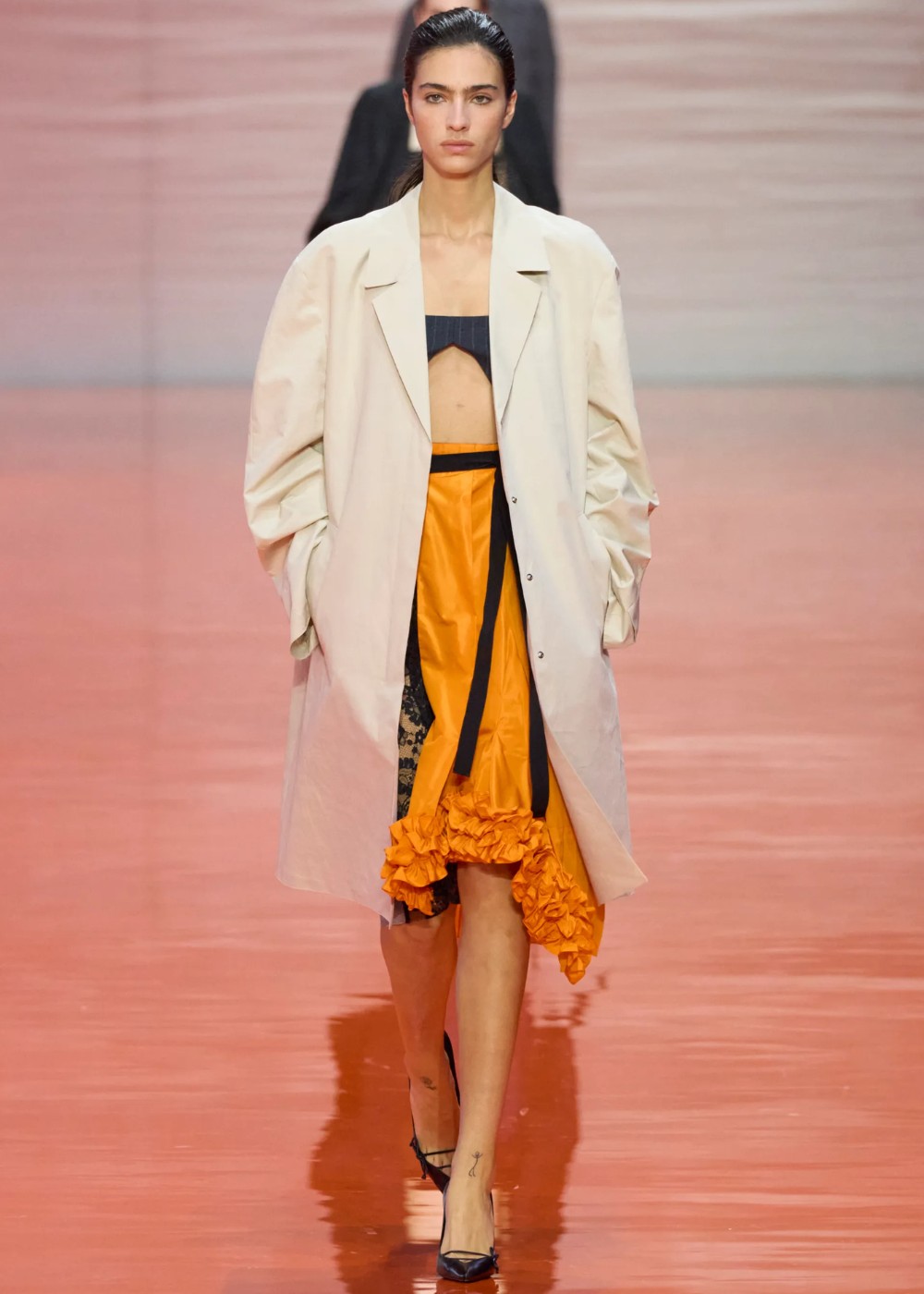 Modelo caminha com postura confiante usando casaco oversized off-white, top cropped preto e saia laranja vibrante.