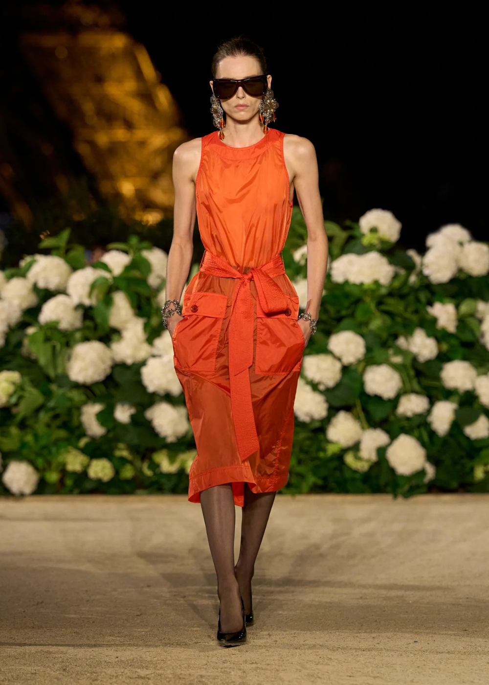 Modelo caminha confiante com vestido midi laranja vibrante em tecido leve, cinto largo e bolsos utilitários.