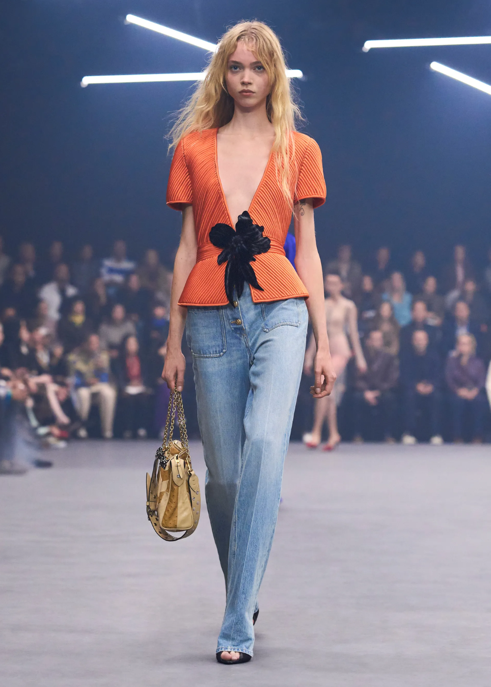 Modelo caminha com postura confiante usando blusa laranja vibrante estruturada com detalhe floral preto e jeans claro.