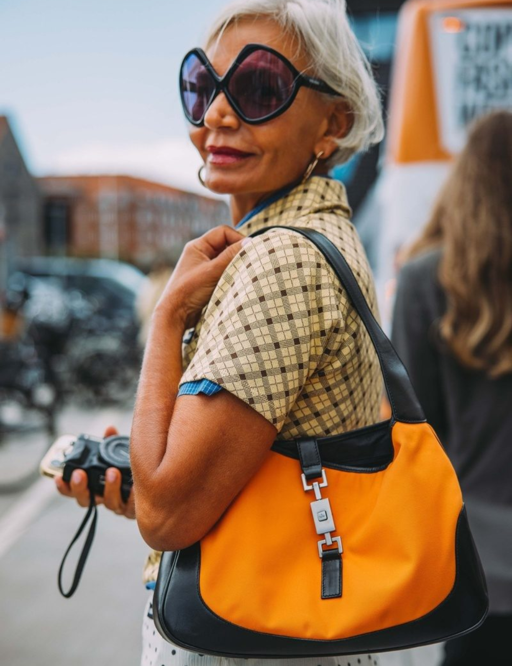 Modelo posa confiante com blazer xadrez bege e marrom, bolsa laranja vibrante e óculos oversized, destacando cores tendência.