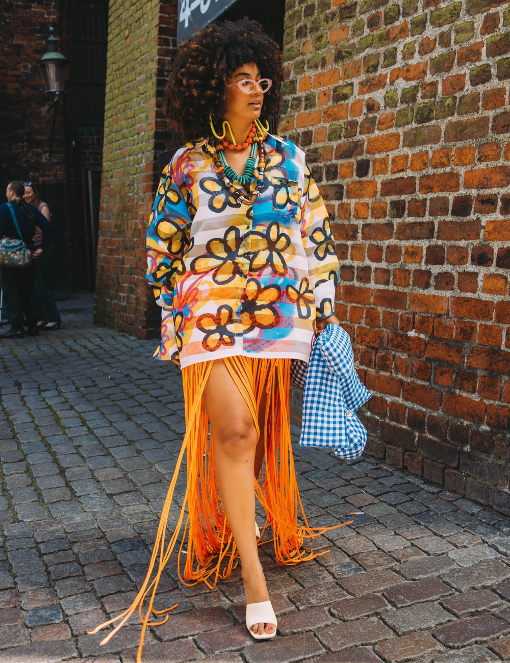 Modelo caminha com camisa ampla estampada em azul, amarelo e laranja, saia de franjas vibrantes e acessórios coloridos.