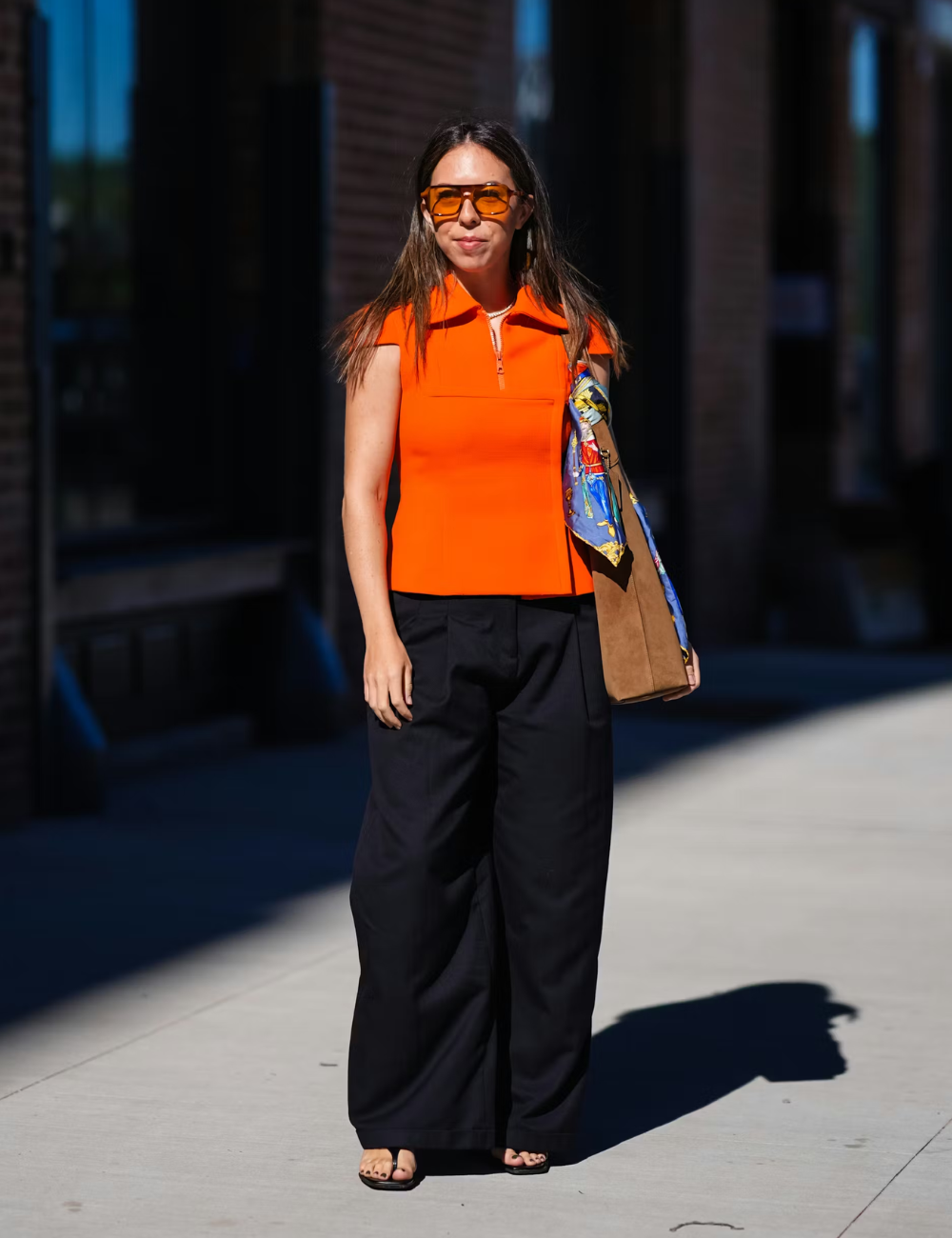 Modelo posa confiante com blusa laranja vibrante de gola estruturada e zíper frontal, calça preta ampla e bolsa camel.