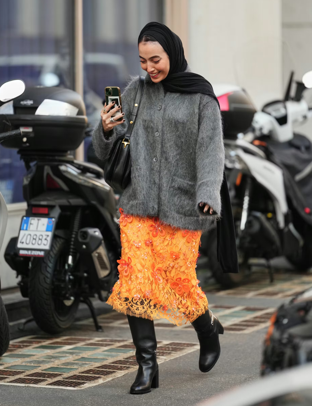 Mulher sorri olhando o celular, veste cardigan cinza felpudo oversized e saia midi laranja vibrante com aplicação floral.