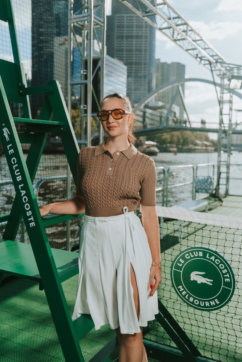 Modelo posa com elegância em ambiente esportivo urbano vestindo polo marrom de tricô e saia Lacoste plissada com fenda