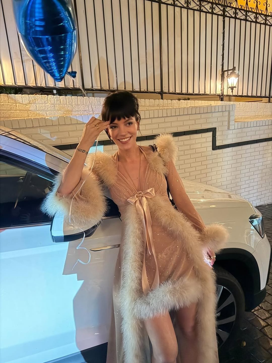 Lily Allen posa sorridente com vestido nude de tule e plumas, laço de cetim e brilho sutil, em clima festivo e elegante.