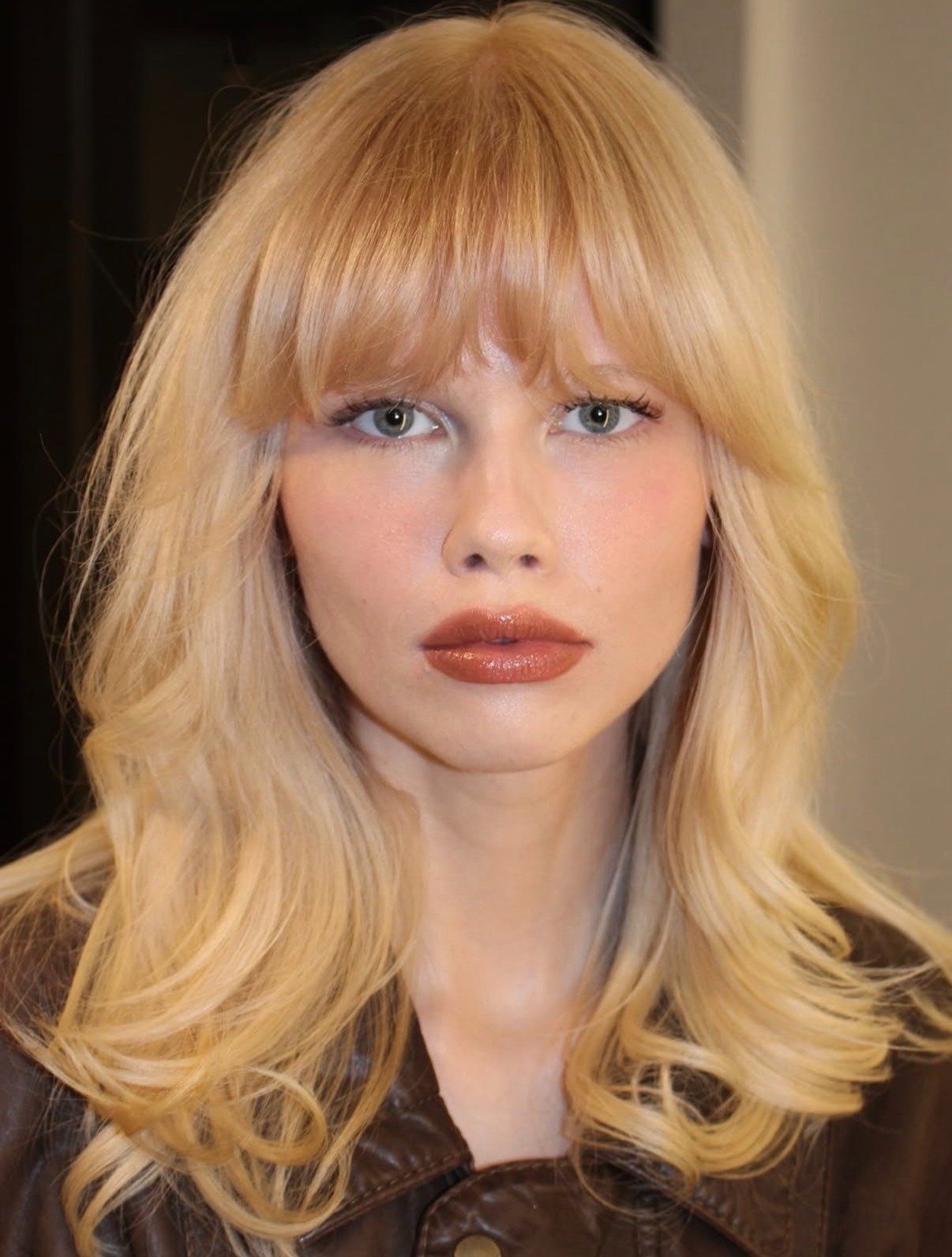 Modelo posa com cabelo loiro de tinta em ondas suaves e franja, usando jaqueta marrom de couro, expressão serena e olhar