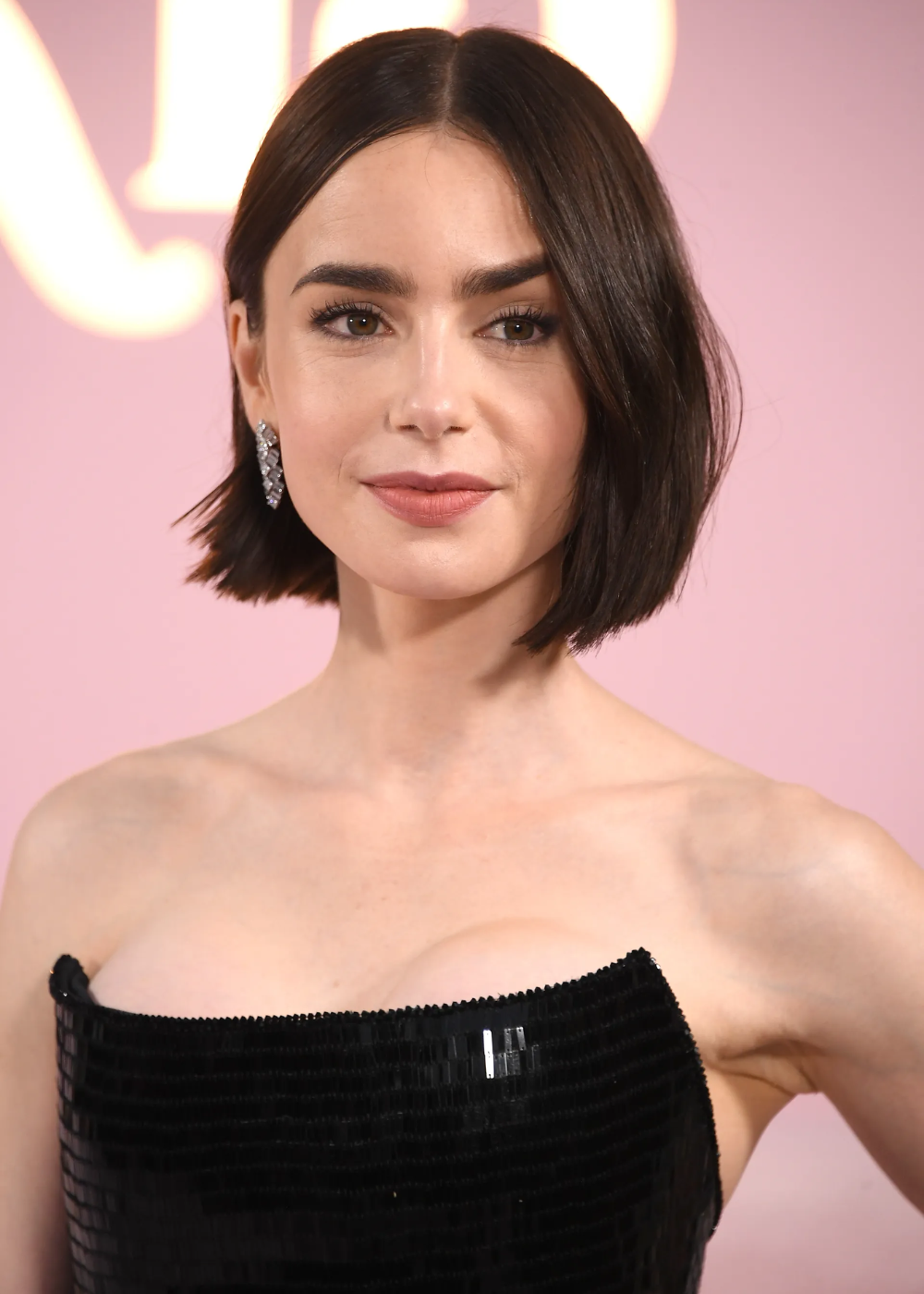 Lily Collins posa com corte bob reto e escuro, vestido tomara que caia preto de paetês e brincos de pedras brilhantes.