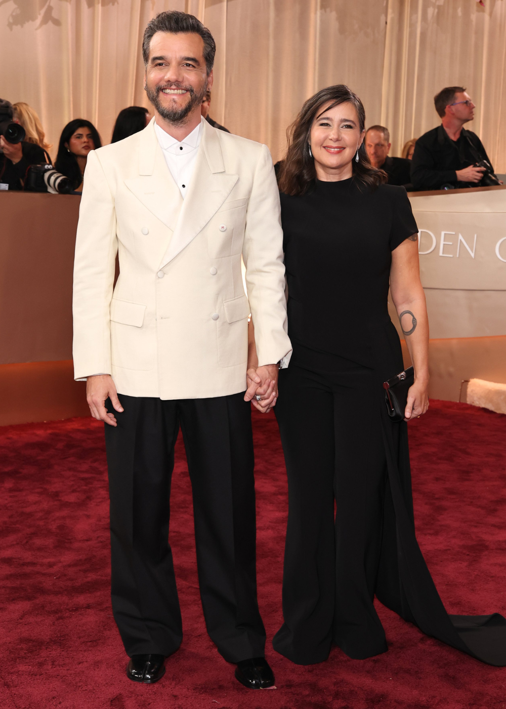 Look de Sandra Delgado. Ela posa sorridente com conjunto preto de manga curta e cauda longa, combinando clutch preta.