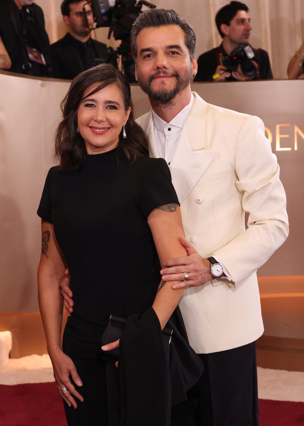 Sandra Delgado veste vestido preto justo de manga curta com clutch e sorriso sereno, posando ao lado de Wagner Moura.