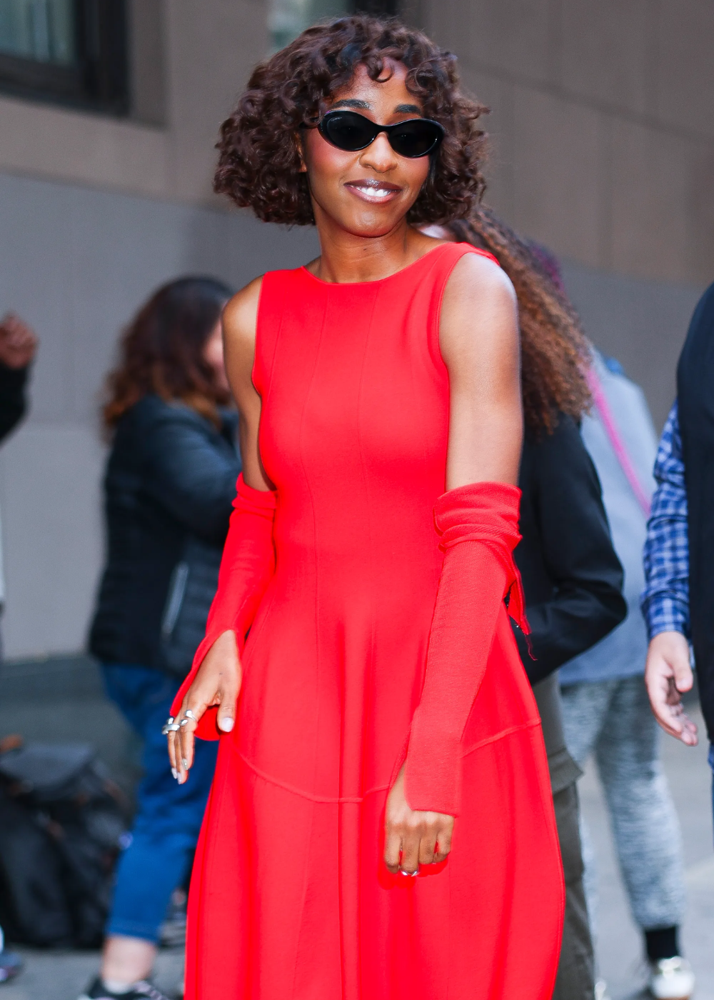Mulher com corte bob cacheado, colete azul marinho ajustado e calça cargo verde, posa sorrindo com bolsa preta em rua urbana.